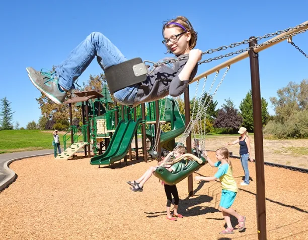 Child with glasses swinging high on a belt swing, while other kids push a swing nearby on a sunny day.