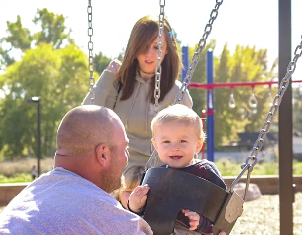 Toddler in a swing smiling at a man, with a woman behind pushing in a sunny playground.