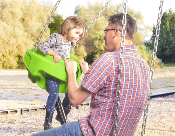 Adult talking to a child on a green bucket swing, surrounded by trees.