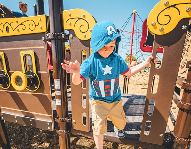 Young child wearing a superhero hoodie walking on a playground bridge with rope net structure in background.