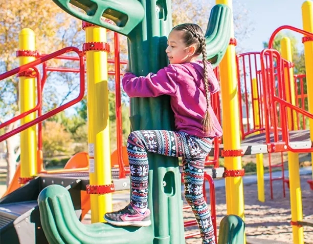 Girl climbing a cactus-shaped structure with colorful playground equipment in the background.