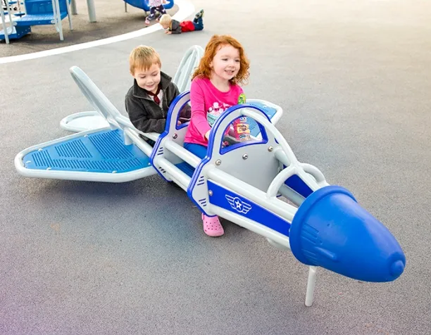 Two children ride a blue and gray airplane-shaped playground rocker on a sunny day.