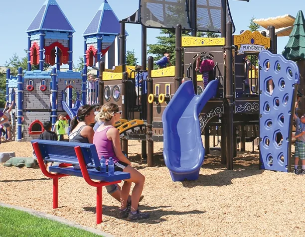 Two women sitting on a red bench watching children play on a castle-themed structure with blue and yellow slides.