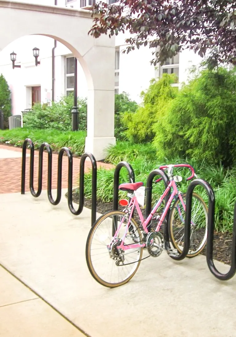 Pink bicycle parked at a black metal bike rack near greenery and building arch.
