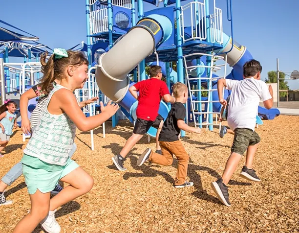 Children running near a large blue playground structure with slides and climbers under a clear sky.