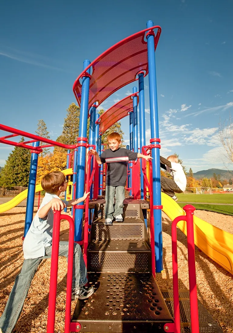 Children climbing a colorful playground structure with red and blue bars under a sunny sky.