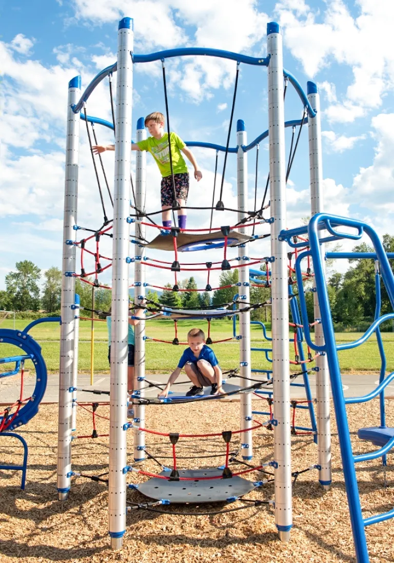 Two children climbing on a tall rope structure in a sunny playground with wood chip surfacing.