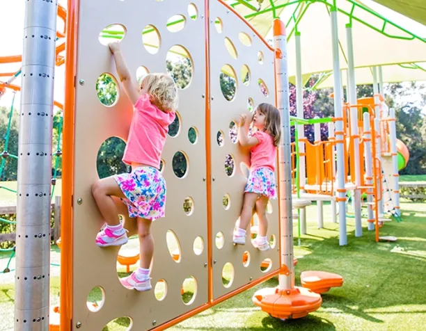 Two young children climbing a beige and orange panel with holes in a brightly lit playground.