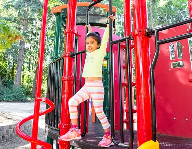 Child hanging from playground bars with red and black climbing structure in a sunny area.