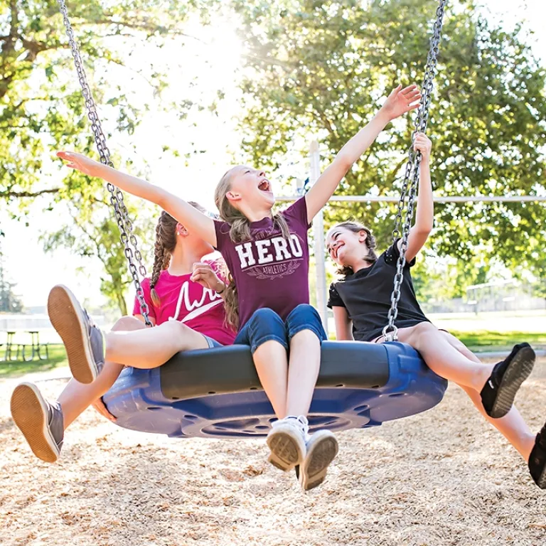 Three girls happily swinging together on a large blue disc swing under sunny skies.
