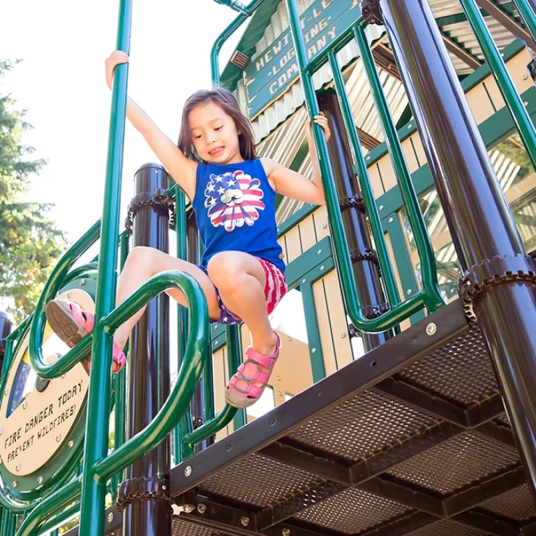 Girl in a blue top playing on green playground bars under bright sunlight.