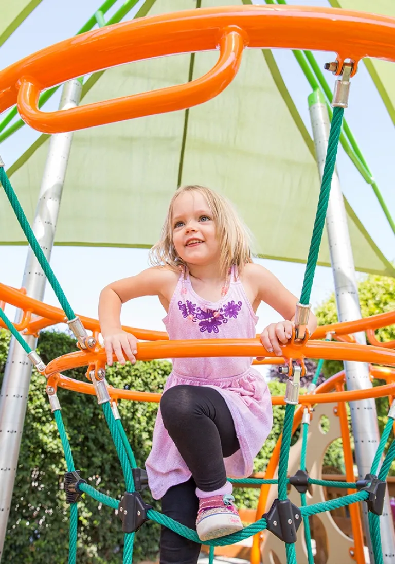 Smiling child climbing on orange and green rope structure under a green shade canopy.