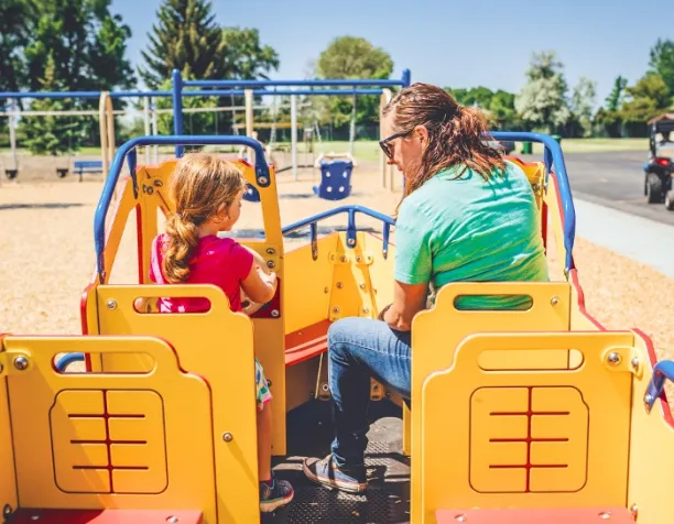 Adult and child sitting on a yellow playground car structure under a sunny sky.