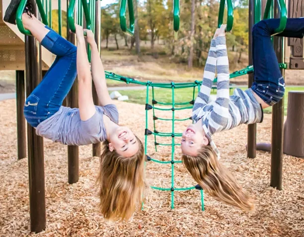 Two girls upside down hanging from green bars on a playground, smiling with wood chips below.