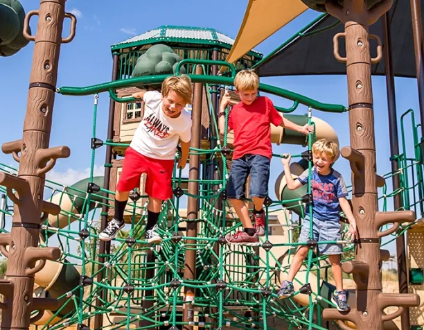 Three children climbing on a green rope net structure under a sunny, clear sky.