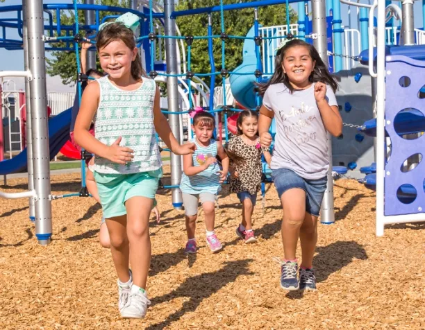 Group of children running on wood chip surfacing near climbing structure in sunny weather.