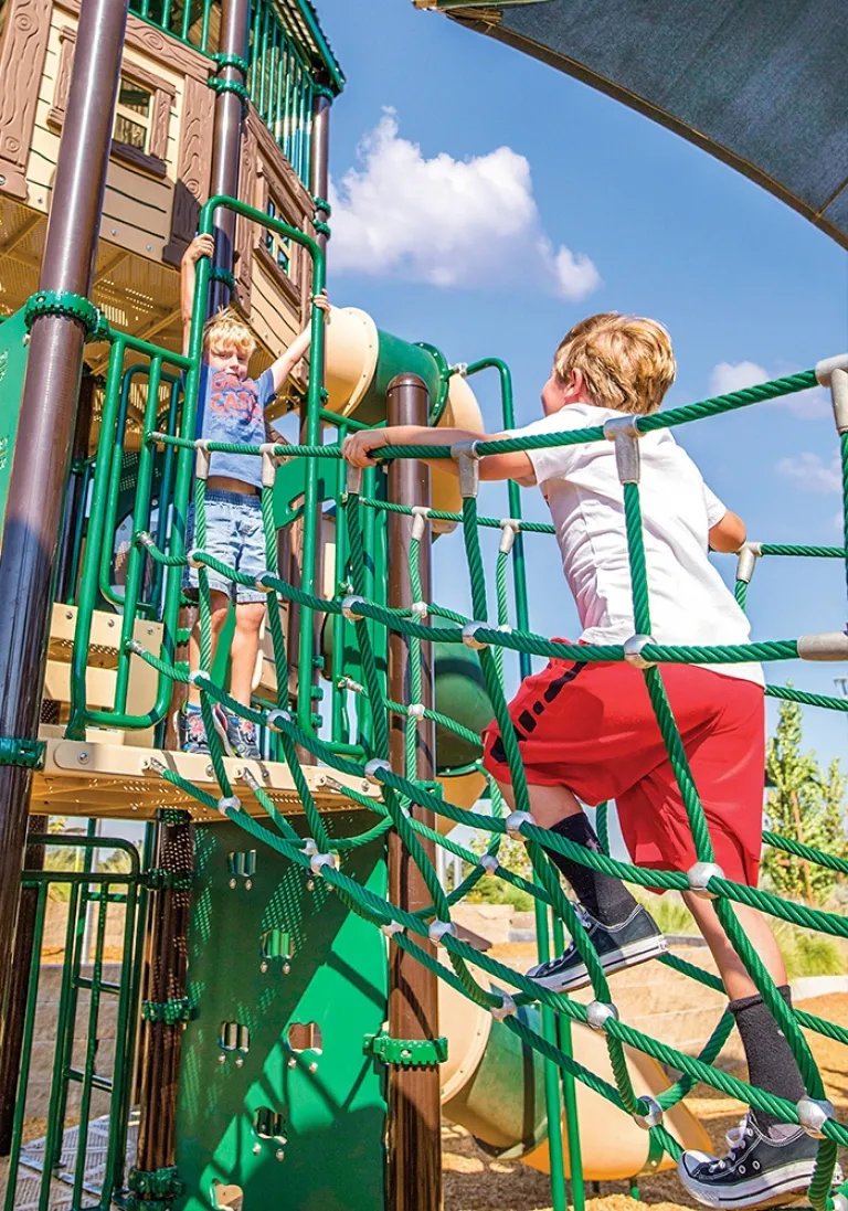 Two children climbing on a green rope net structure under a sunny sky.
