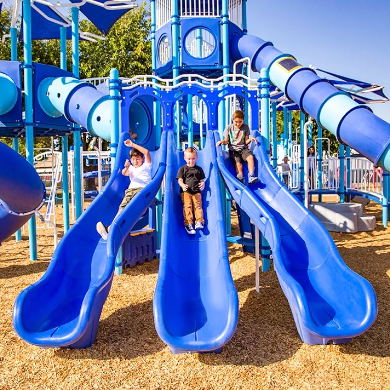 Three children sliding down parallel blue slides under a sunny sky.