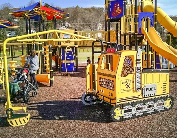 Playground featuring a yellow construction-themed play structure and overhead climber with a parent pushing a stroller.