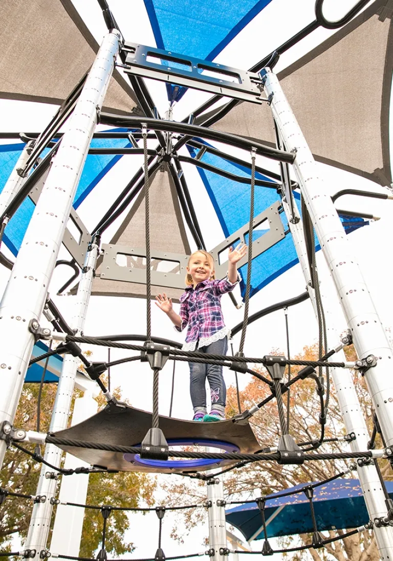 Smiling child standing on a platform in a rope net structure under blue and gray shade canopies.