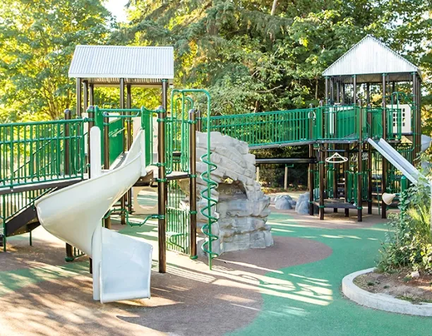 Playground with green structures, tube and spiral slides, rock wall under sunny trees.