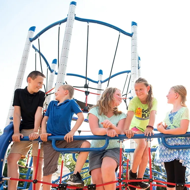 Group of happy children sitting and talking on a rope climbing structure under a sunny sky.