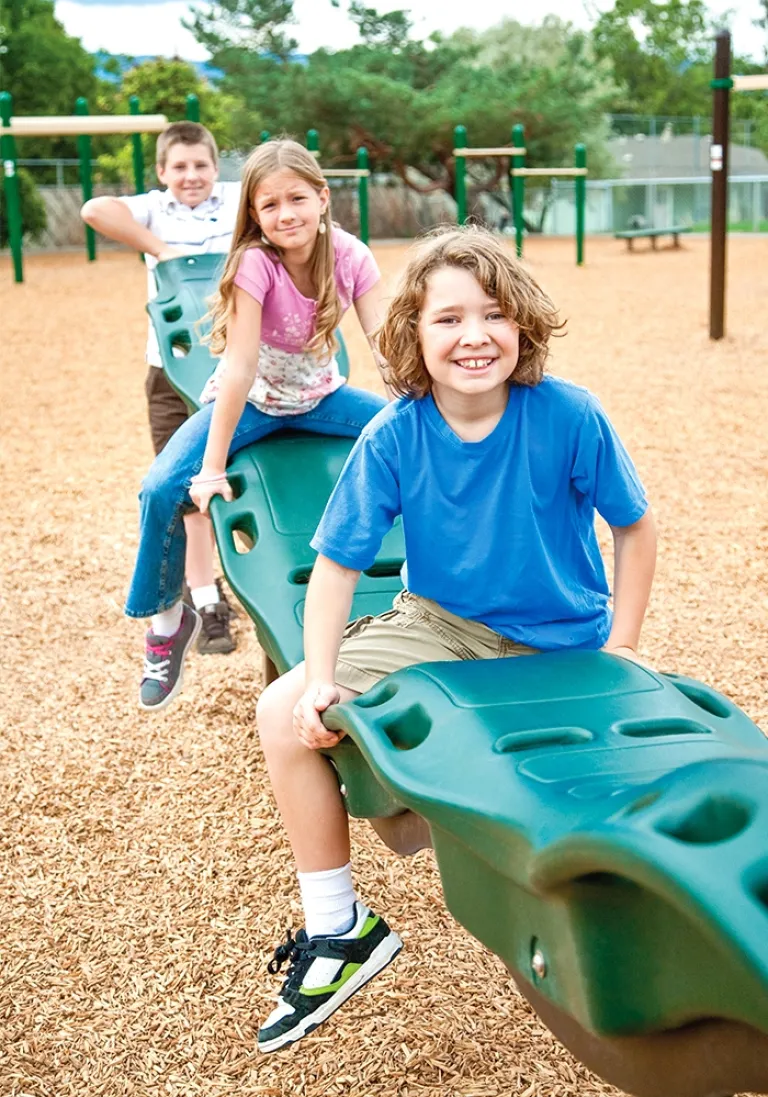 Three children smiling while playing on a green balance beam structure on wood chips.