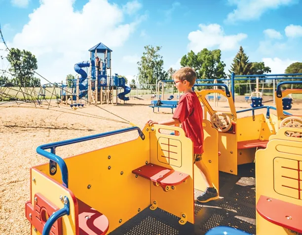 Boy in red shirt playing on yellow and blue playground structure with a rope net and slide in the background.