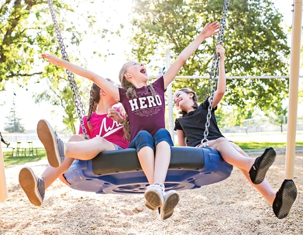 Three laughing girls on a large blue circular swing under sunny trees.