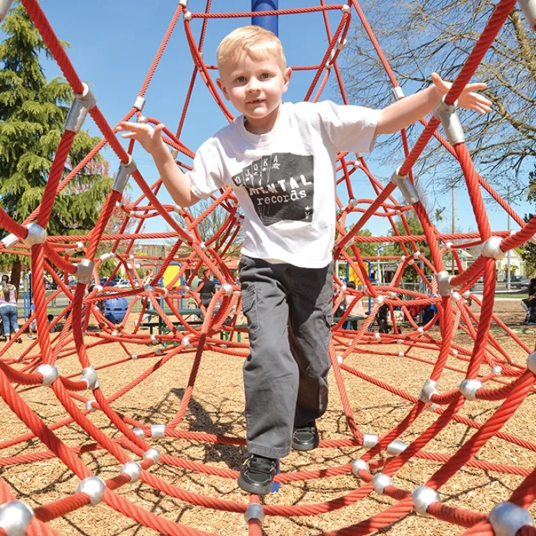 Smiling child climbing on a red rope net in a sunny park.