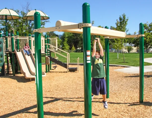 Boy swinging on overhead bars, another child slides nearby, bright sunny day, green and beige playground equipment.