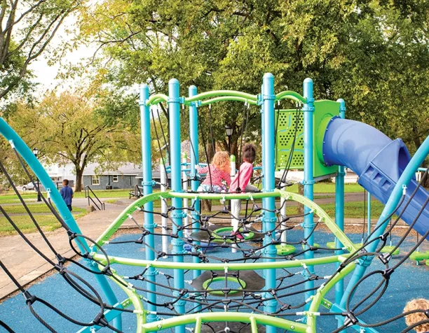 Children seated on a climbing structure with a blue slide in a tree-lined area.