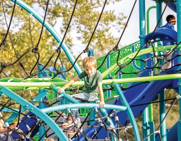 Children climbing on a colorful rope net structure with slides in a sunny park.