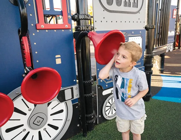 Young boy listening to a talk tube on a train-themed playground structure.
