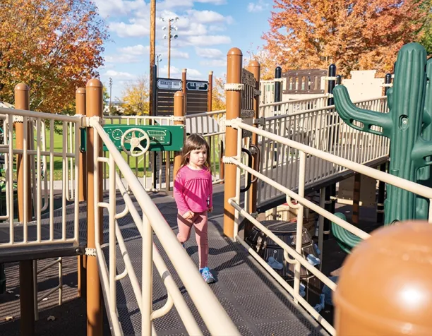 Child in a pink outfit walking on a play structure bridge with autumn trees in the background.