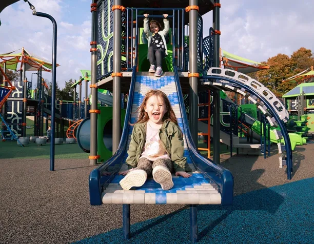 Two children sliding down a wide blue slide in a sunny playground.