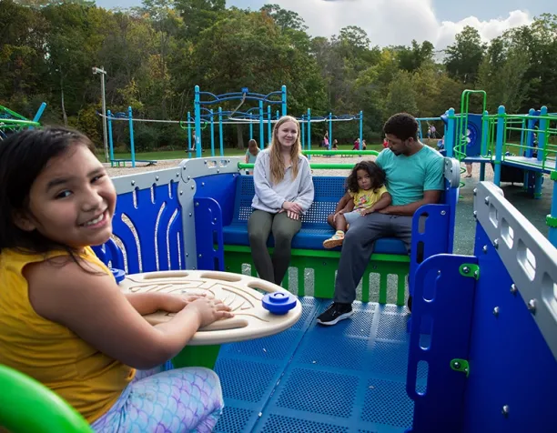 Smiling girl in yellow seated at a play structure with family in the background.