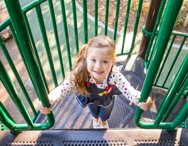 Smiling child in polka dot shirt climbing green playground stairs.