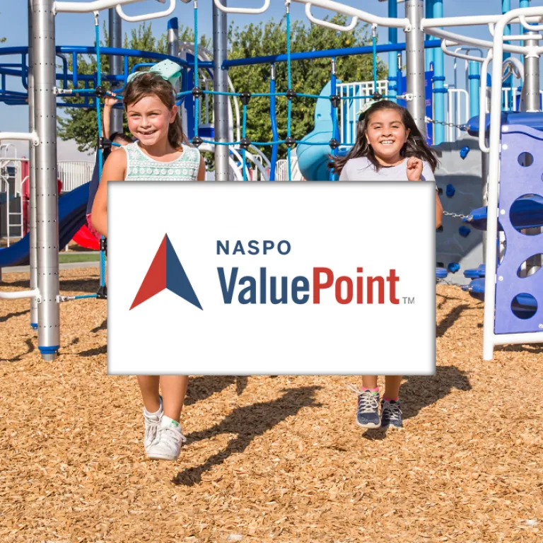 Two smiling girls running on wood chips under a blue and white climbing structure.
