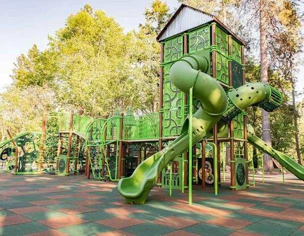 Green playground structure with spiral slide and climbing features surrounded by trees.