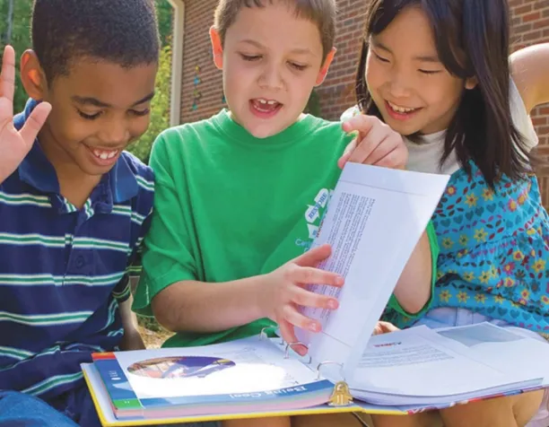 Three children sitting together, looking at a book, and smiling outside.