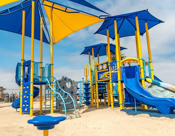 Colorful playground with yellow and blue slides, climbing structures, and shade canopies in a sunny setting.