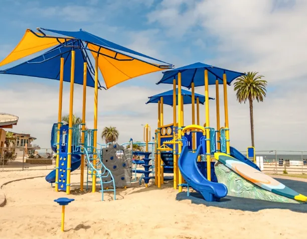 Playground with blue and yellow shade structures, slides, and climbing tower on sandy beach.