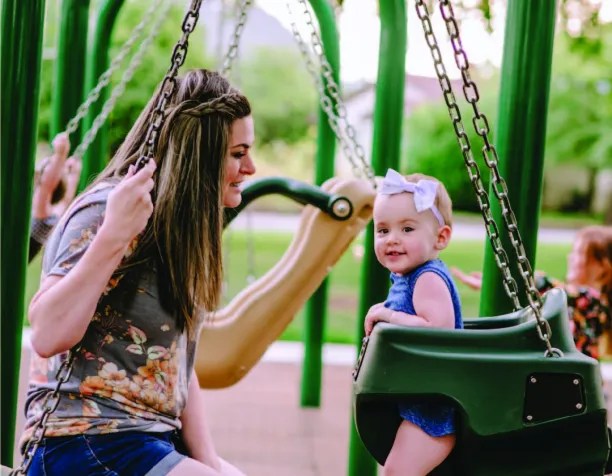 Woman and toddler facing each other on adjacent swings in a playground with green supports.