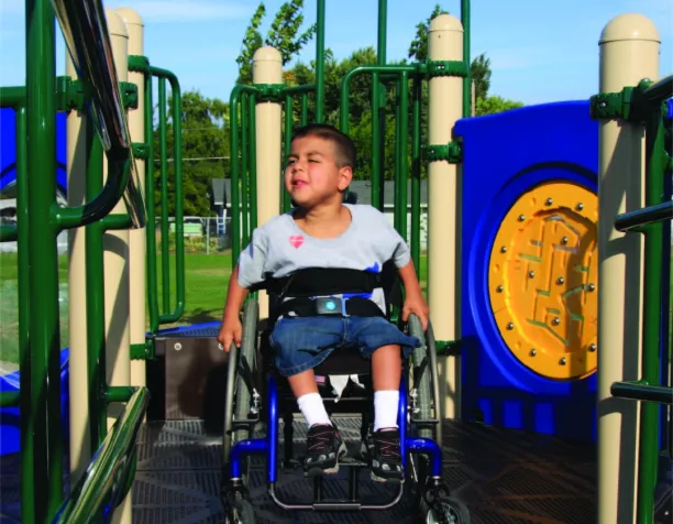 Child in a wheelchair enjoying an accessible playground with ramps and colorful panels.
