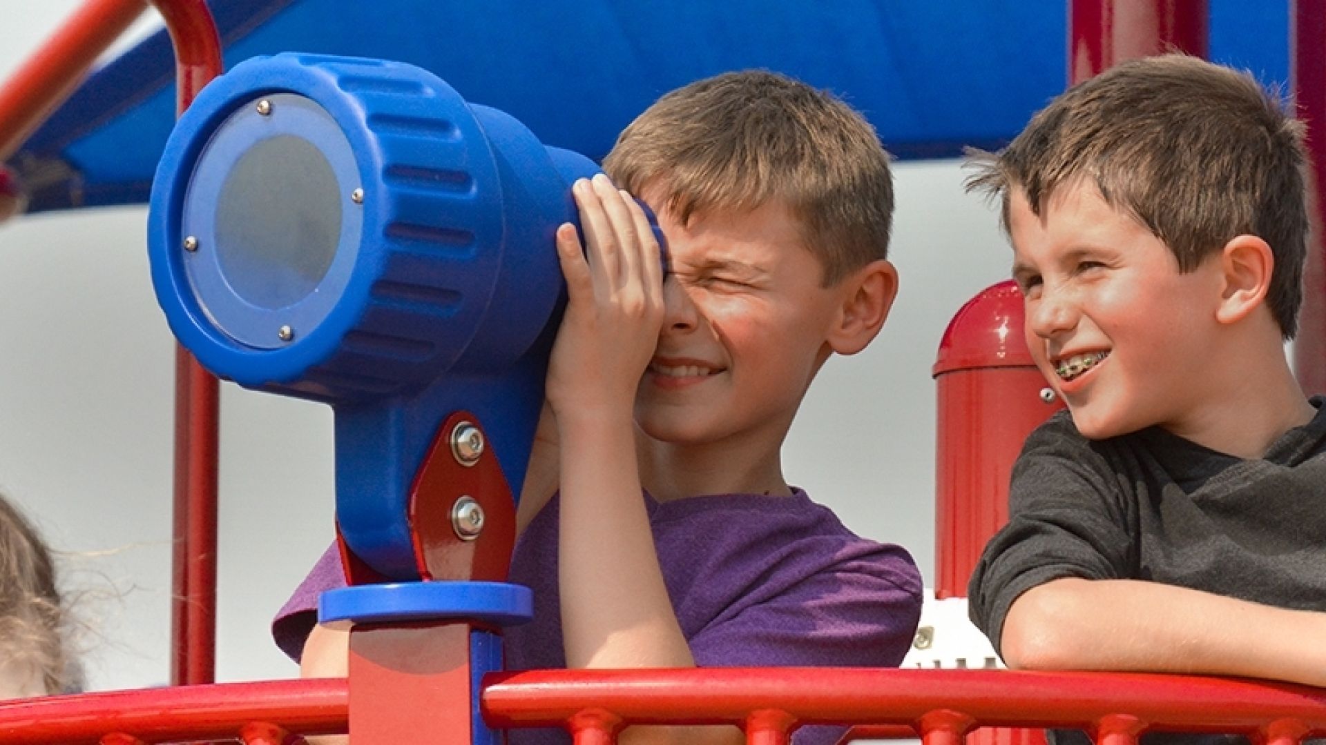Two boys on a play structure using a blue telescope under a red and blue shade.