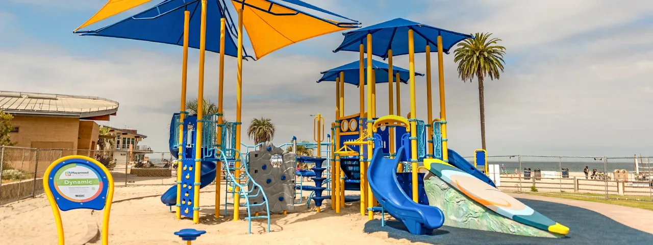 Playground with blue and yellow slides and shade structures near a beach.